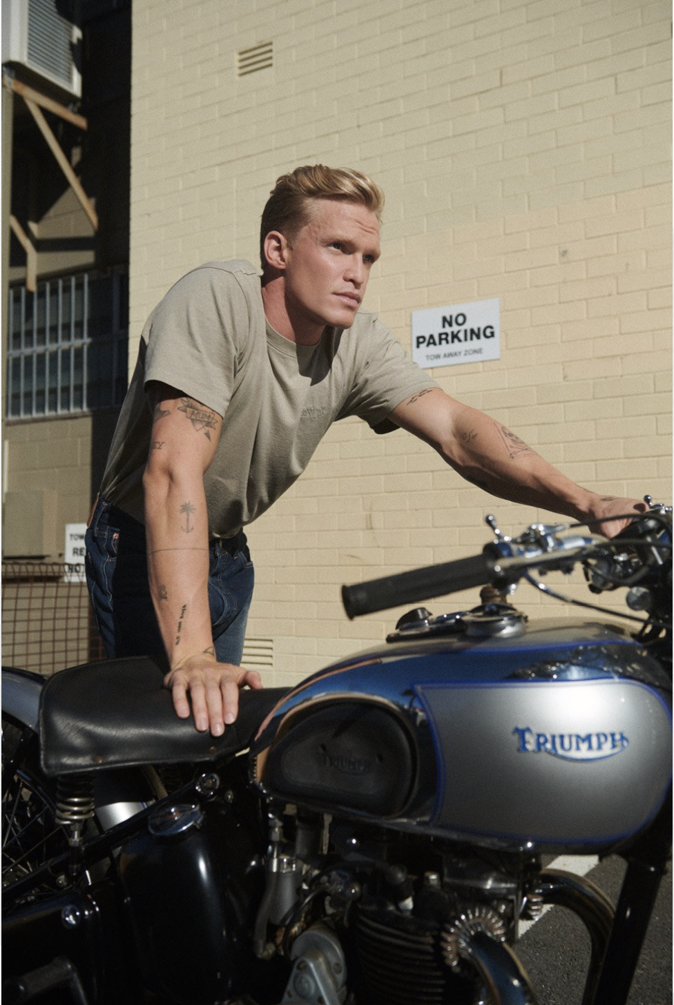 Adult male leaning on a blue and silver Triumph motorcycle in front of a tan brick building with a 'No Parking' sign.