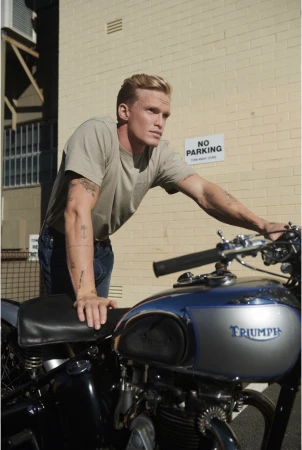 Adult male leaning on a blue and silver Triumph motorcycle in front of a tan brick building with a 'No Parking' sign.