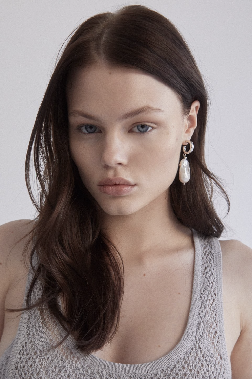 Close-up portrait of a woman with long brown hair wearing a grey tank top and a statement earring with a pearl drop.