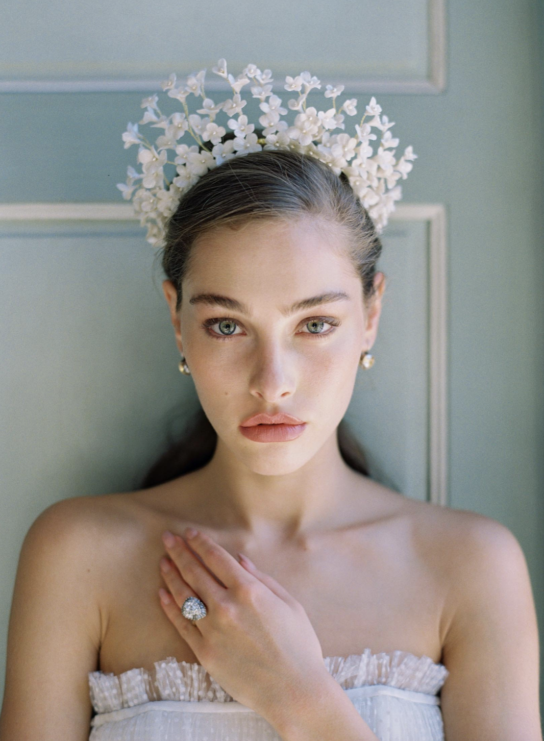 Elegant portrait of a woman with floral headpiece and jewelry against a light blue panel wall in soft, diffused lighting.
