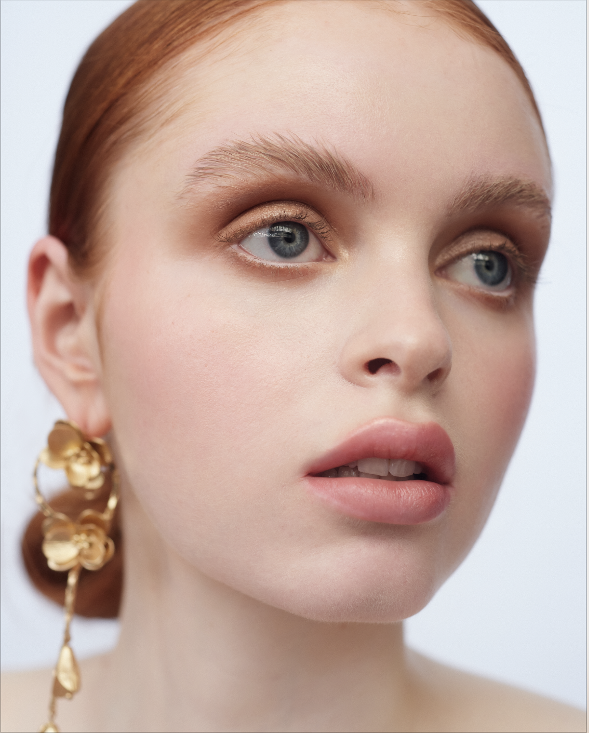 Close-up portrait of a redhead woman with dramatic makeup and gold flower earrings against a clean white backdrop in a beauty editorial.