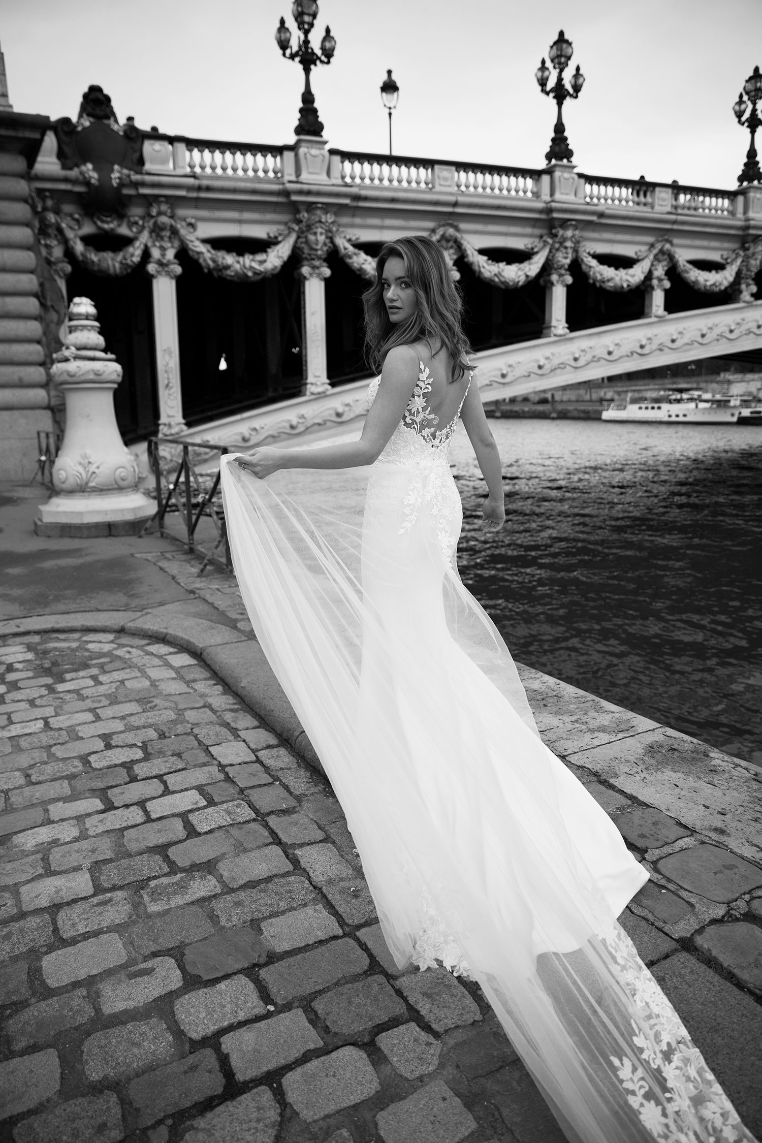Woman in wedding dress poses on cobblestone street by Seine River, Paris, with bridge in background. Back view with flowing fabric.