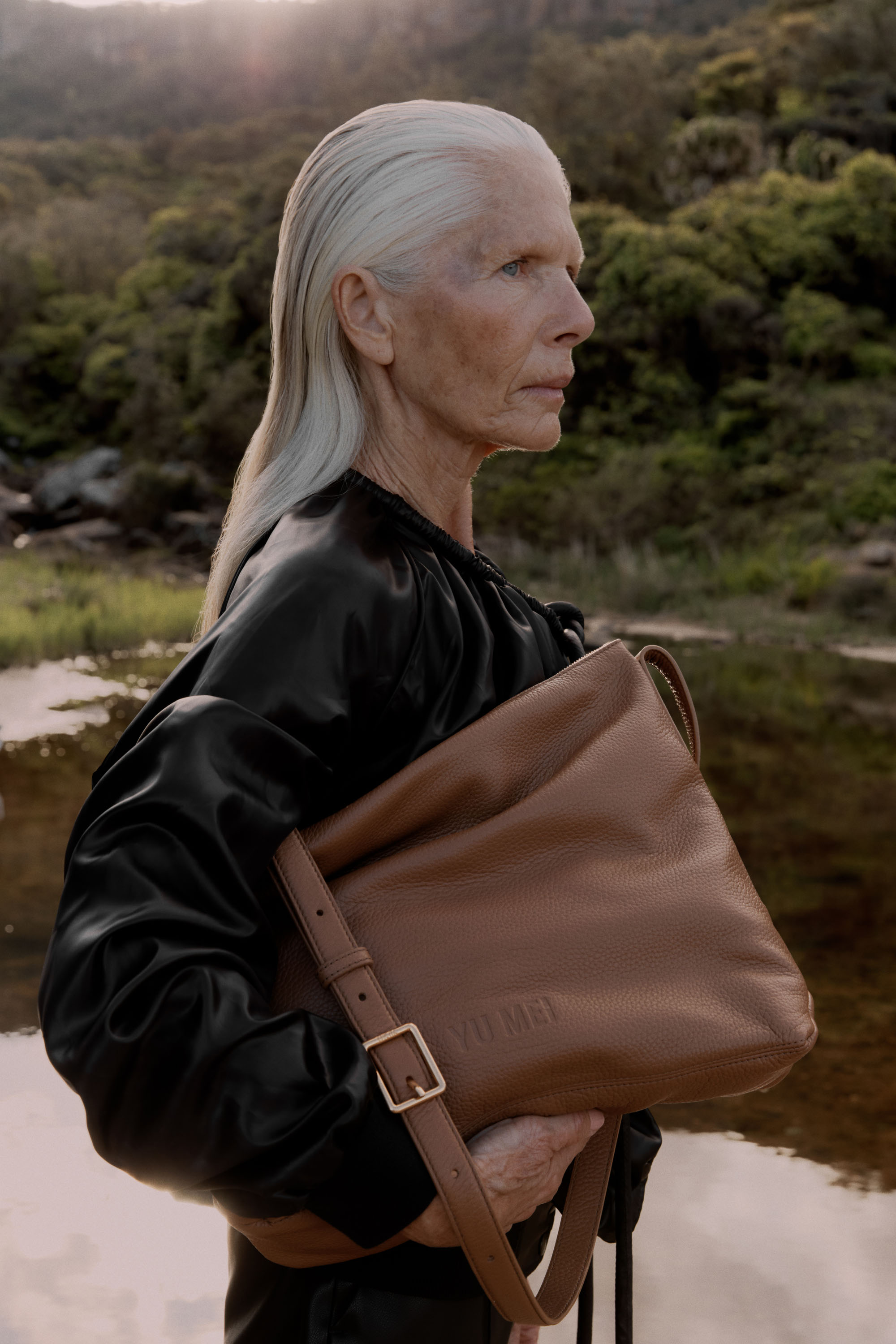 Mature woman with silver hair stands outdoors in a black outfit holding a large brown leather handbag in a tranquil landscape.