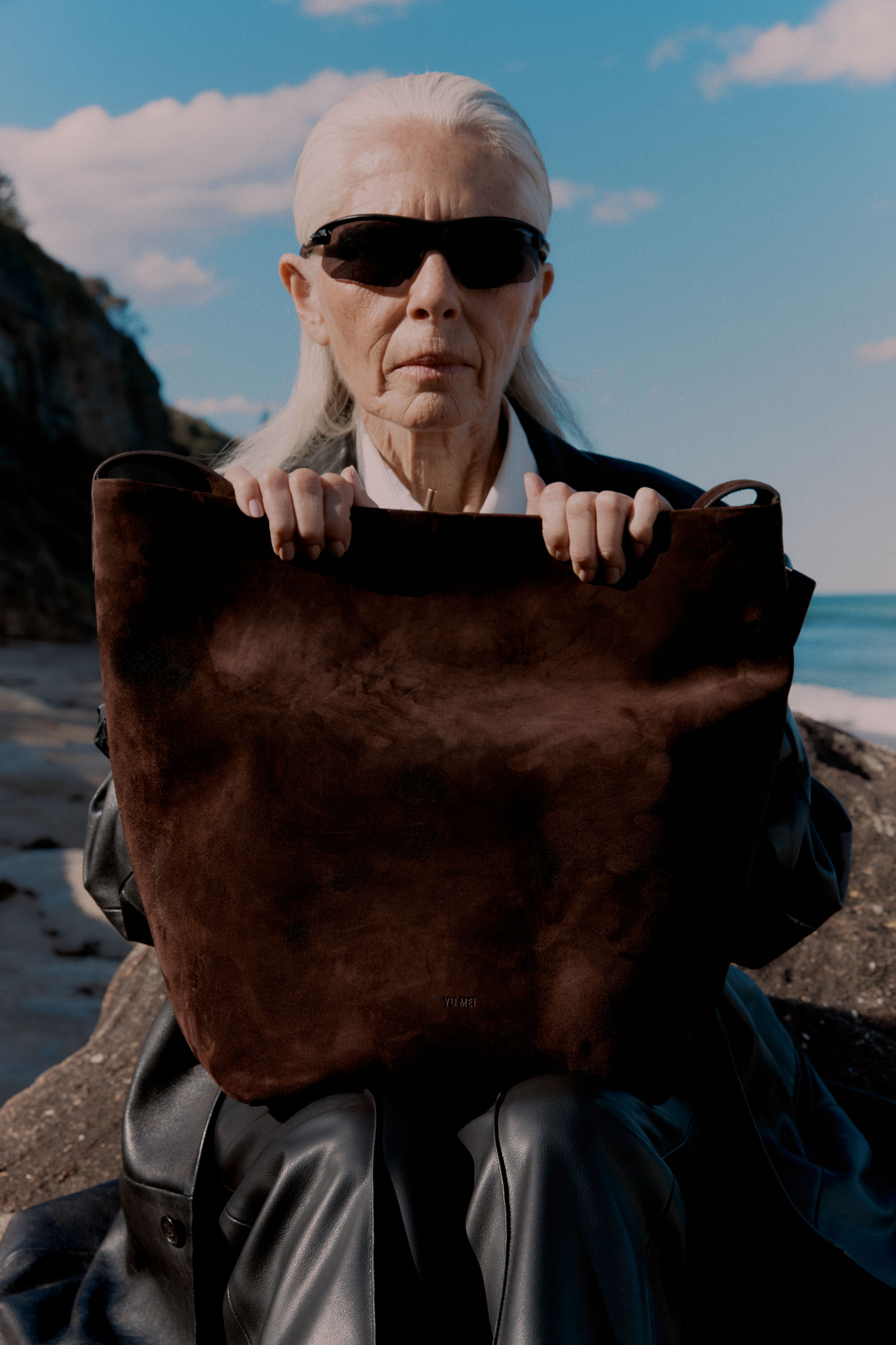 Elderly woman with long white hair poses on a rocky beach with a large brown suede tote bag, wearing sunglasses and a black leather jacket.