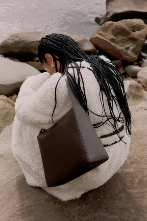 Woman in white fluffy coat with long black braided hair holding a brown leather shoulder bag on a rocky coast