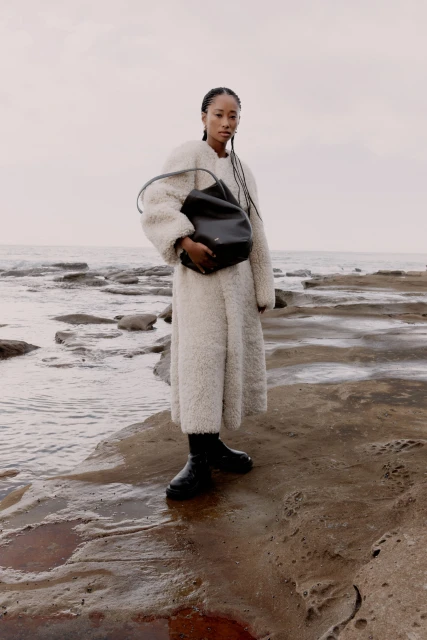 A woman in a white coat holds a black bag on a rocky beach with the ocean and overcast sky in the background, creating a stylish editorial shot.