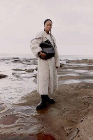 A woman in a white coat holds a black bag on a rocky beach with the ocean and overcast sky in the background, creating a stylish editorial shot.