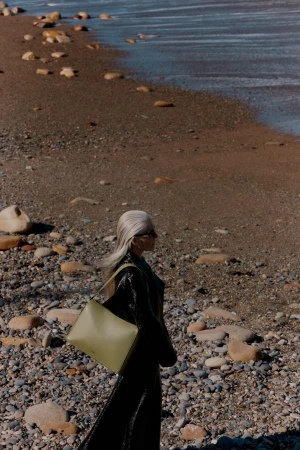 Woman with long silver hair wearing dark coat carrying an olive green shoulder bag walking along a pebble beach by the ocean.