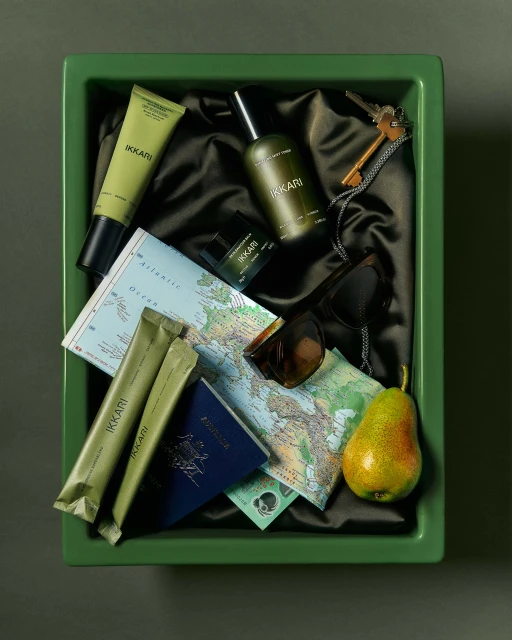 Overhead shot of travel essentials in green container: beauty products, passport, maps, keys, sunglasses, currency, and a pear.