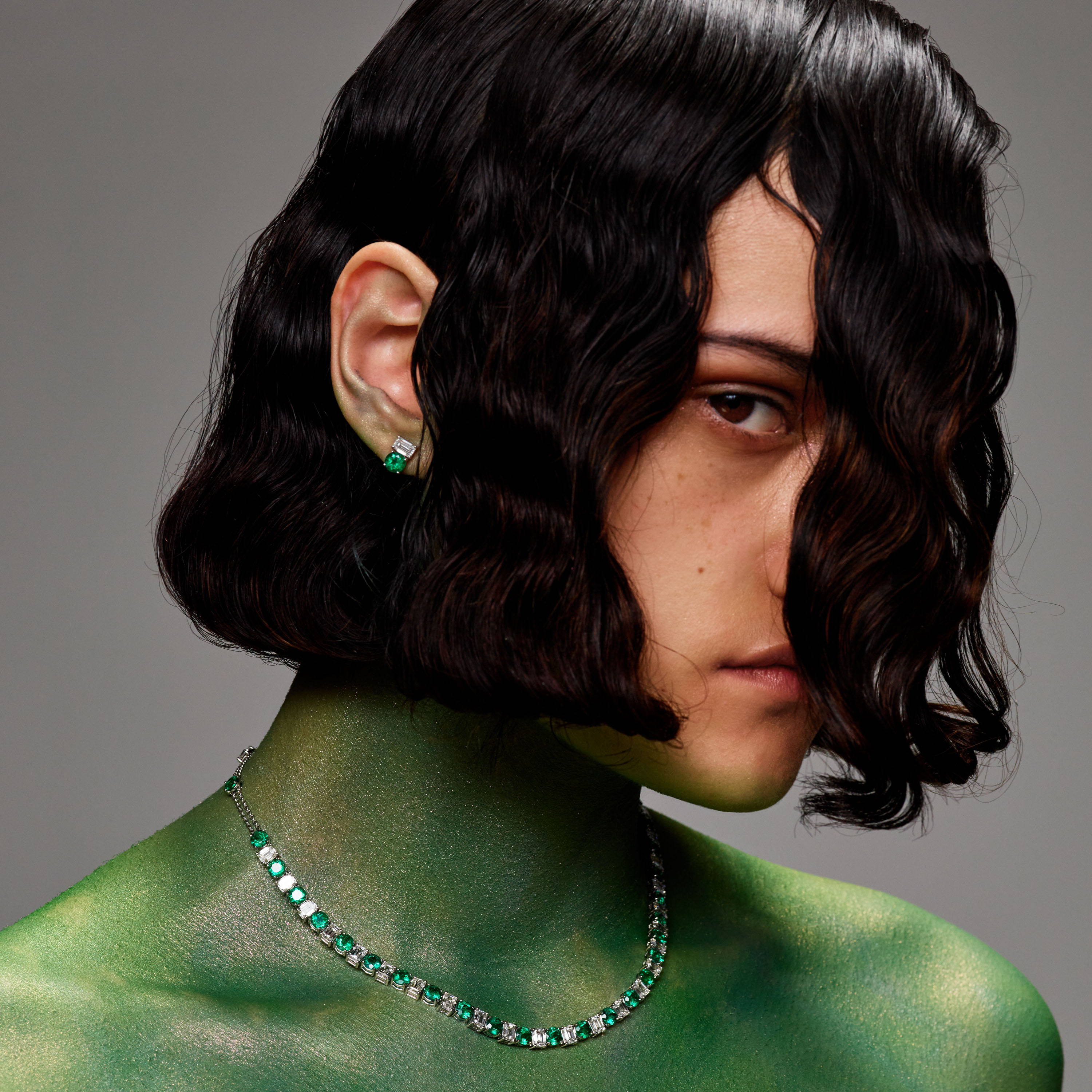 Close-up shot of a brunette woman wearing a green body paint and a diamond and emerald necklace and earring set against gray background.