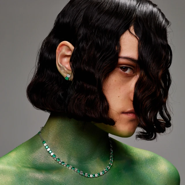 Close-up shot of a brunette woman wearing a green body paint and a diamond and emerald necklace and earring set against gray background.