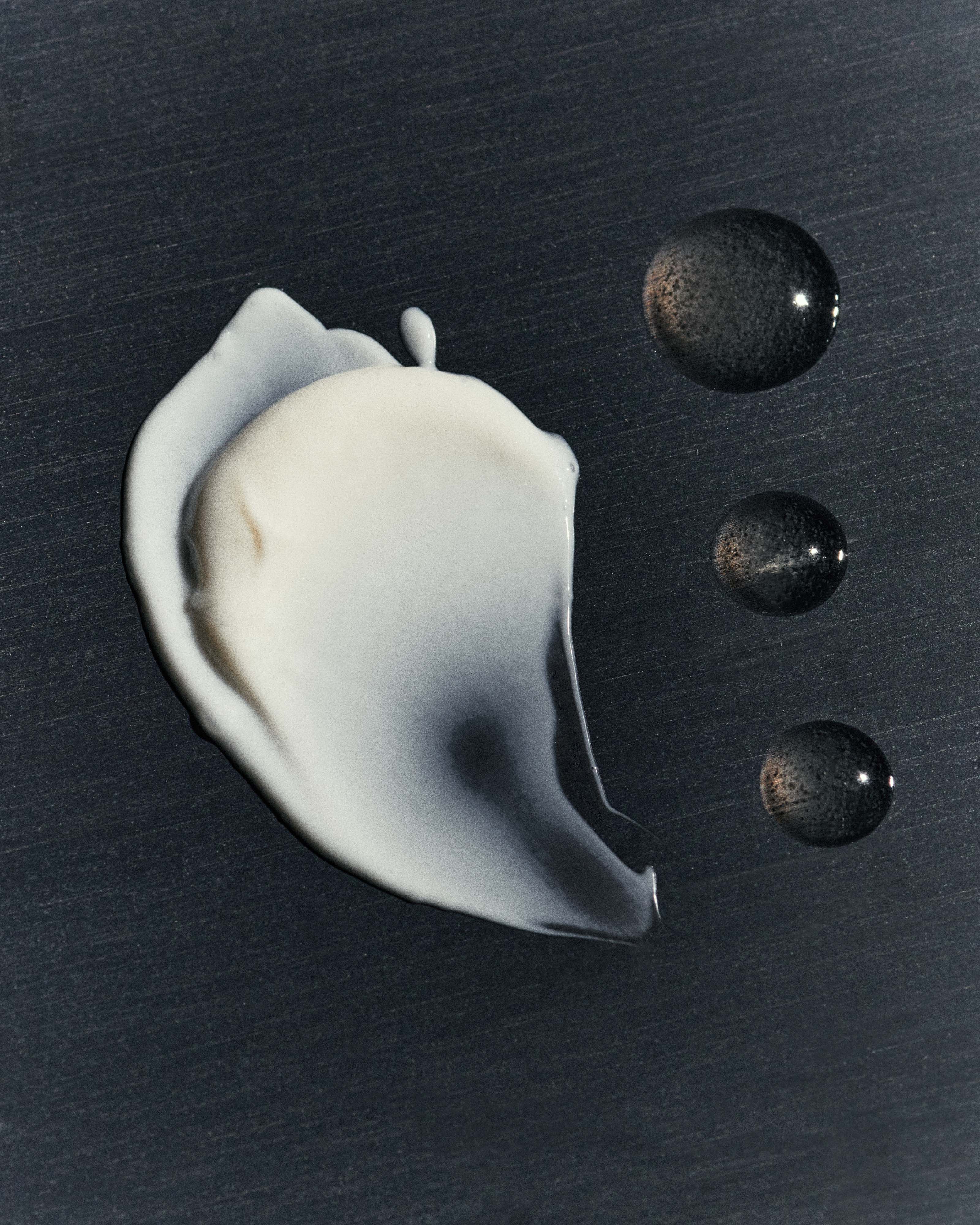 Close-up studio shot of a smear of white beauty cream with three adjacent dark liquid drops against a dark grey background.