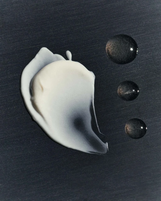 Close-up studio shot of a smear of white beauty cream with three adjacent dark liquid drops against a dark grey background.