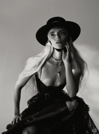 Monochrome portrait of a woman with long blonde hair wearing a black hat and dark dress against a plain background
