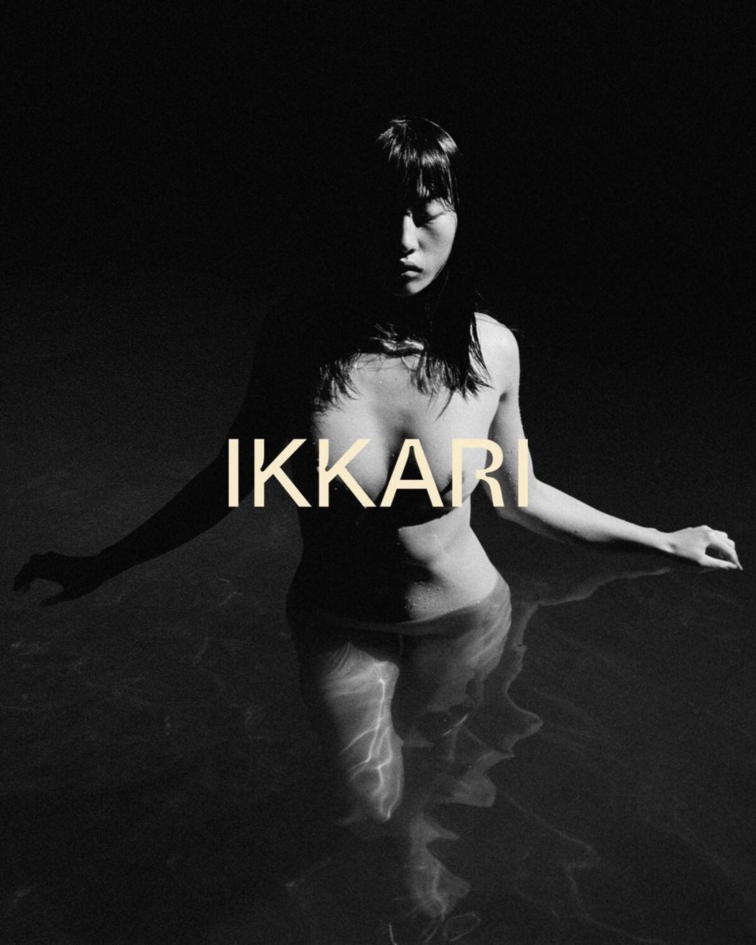 Monochrome portrait of a woman standing in water, her upper body and face partially shadowed, with the word 'IKKARI' overlaid.