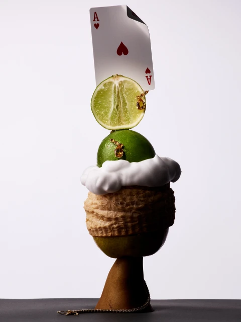 Whimsical still life composition features stacked fruits, a playing card, and gold leaf against a gradient white and gray background.