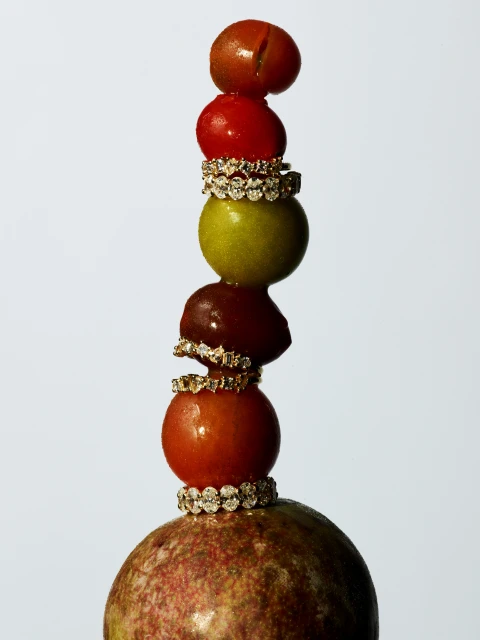 A stacked arrangement of passion fruit, tomatoes, and olive held together by diamond rings on a white background.