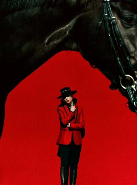 Fashion shot of a woman in a red jacket and hat standing next to a black horse against a red backdrop in a studio setting.
