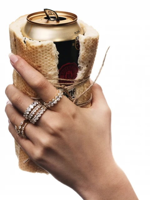 A hand holds a beer can wrapped in bread and tied with twine. The hand wears three rings, and the background is white.