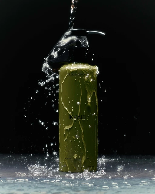 A product shot of a green pump bottle of shampoo being sprayed with water against a dark backdrop, creating splashes of water.
