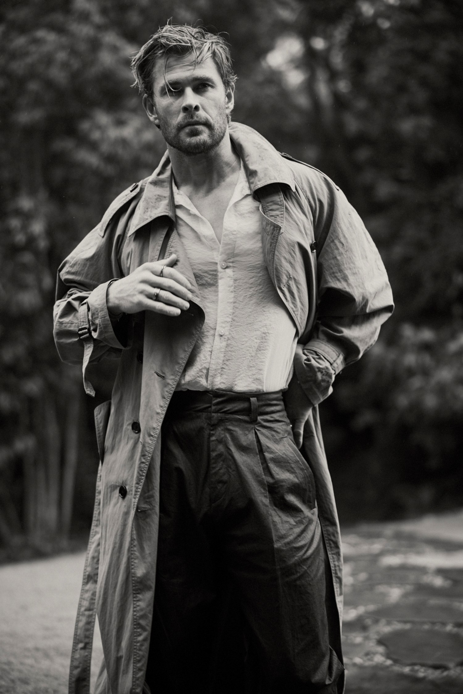 Black and white portrait of Chris Hemsworth in a trench coat against an outdoor background with foliage and soft, diffused lighting.
