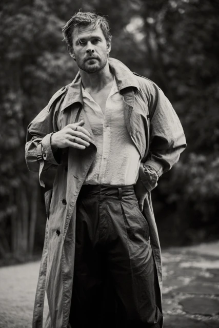 Black and white portrait of Chris Hemsworth in a trench coat against an outdoor background with foliage and soft, diffused lighting.