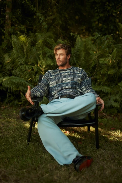 Chris Hemsworth sits in a black chair on grass in a wooded setting, wearing a patterned sweater and light blue pants, looking away from the camera.