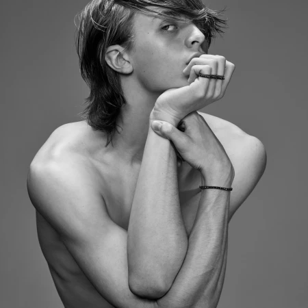 Portrait of a shirtless young man with dark tousled hair resting his chin on his hands against a seamless grey backdrop