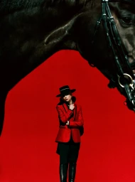 Model in red blazer and black hat standing beneath a large horse head against a vibrant red backdrop