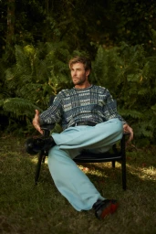 Chris Hemsworth seated in an outdoor black chair wearing a patterned blue knit sweater and light blue trousers against a lush forest backdrop.