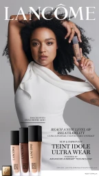 A woman in a white one-shoulder dress holding a Lancôme foundation bottle against a grey studio background