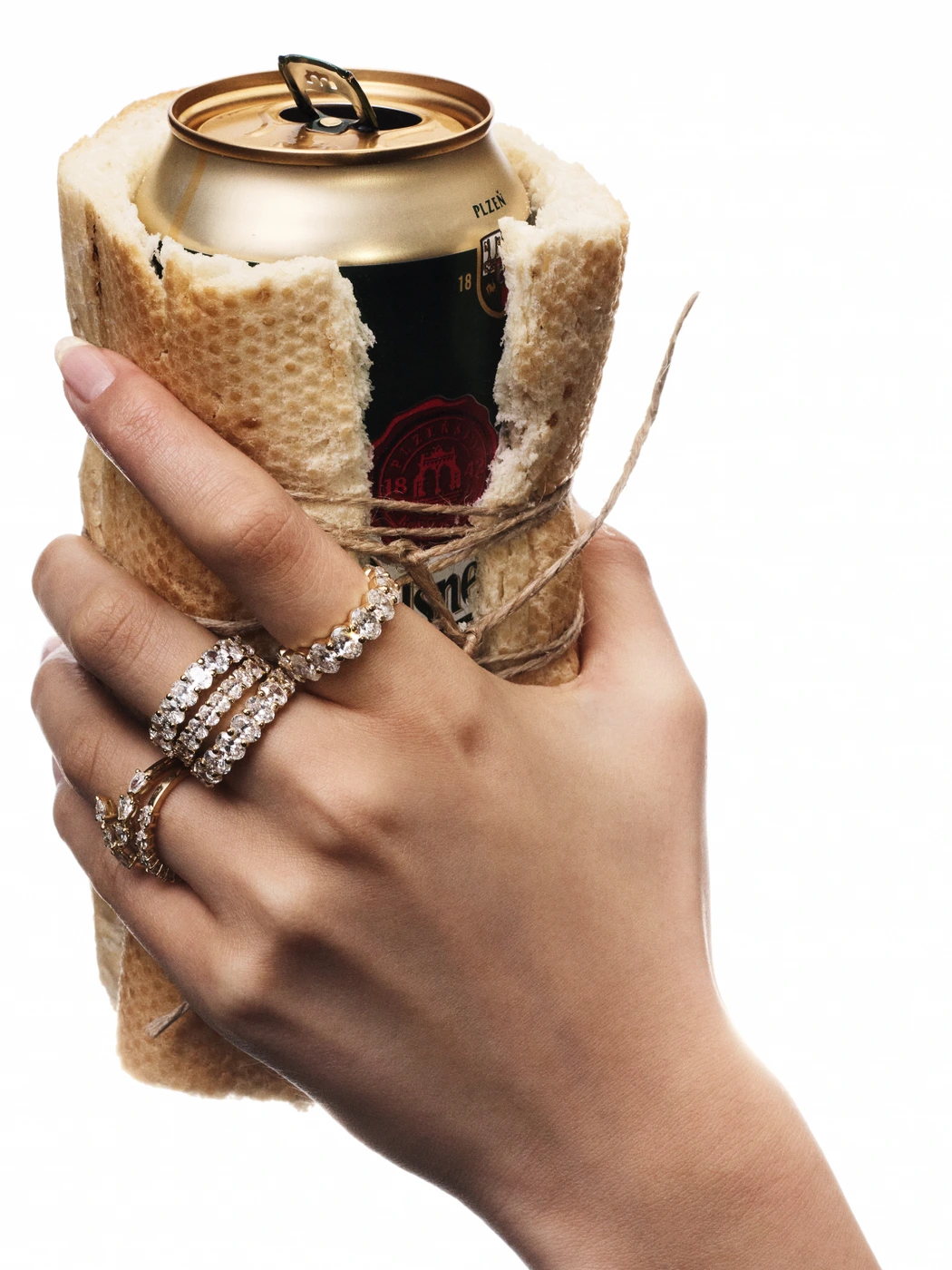 Hand wearing diamond rings holding a beverage can wrapped in a baguette tied with string against a white background