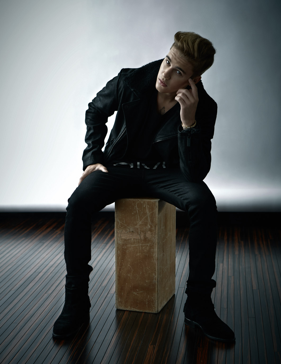 Adult male posing indoors. Man with blond hair wearing a black jacket and pants sitting on a wooden stool in a studio setting.