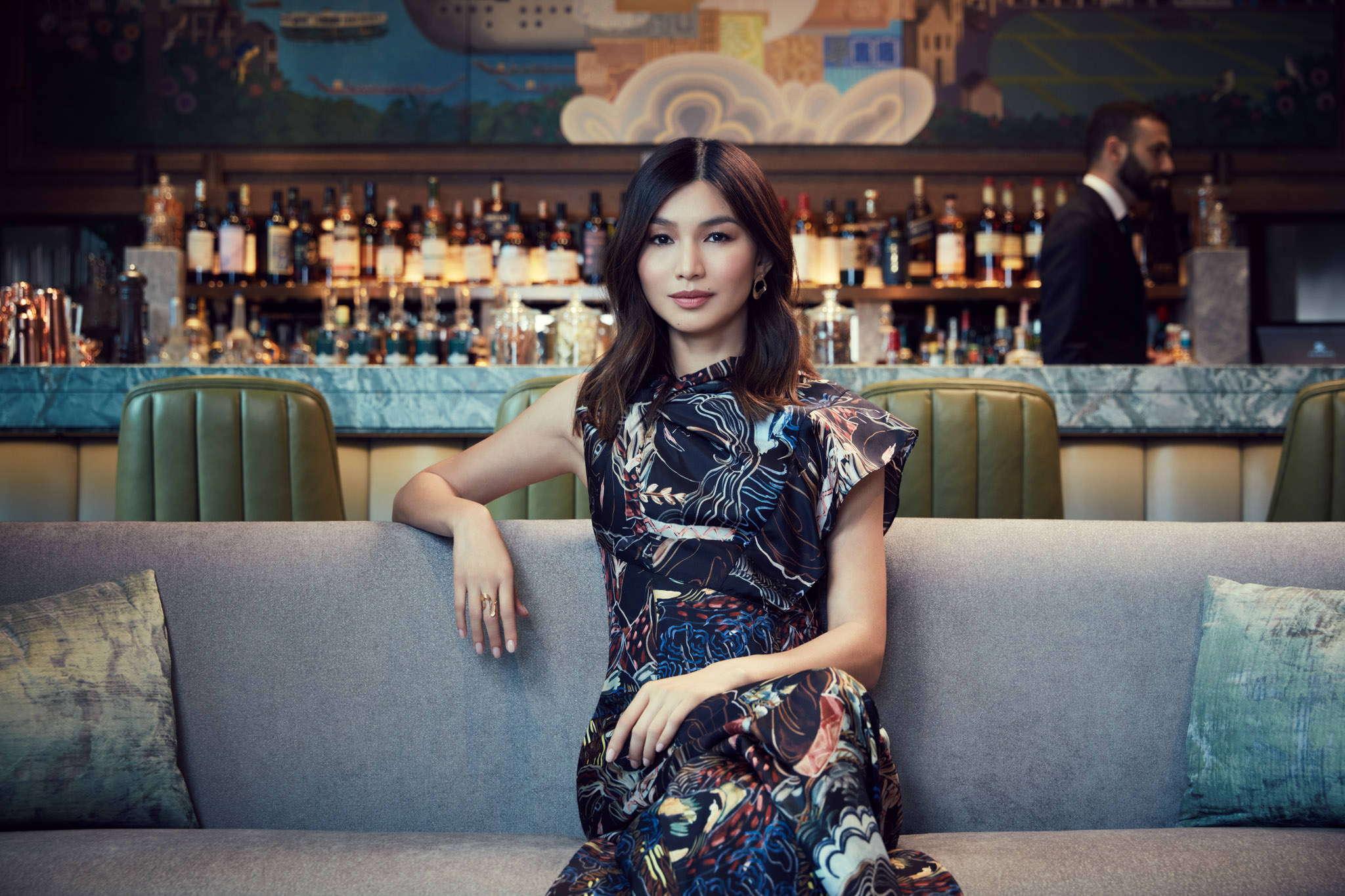 Adult female posing in patterned sleeveless dress on gray sofa indoors at bar with leather chairs and mural in the background.