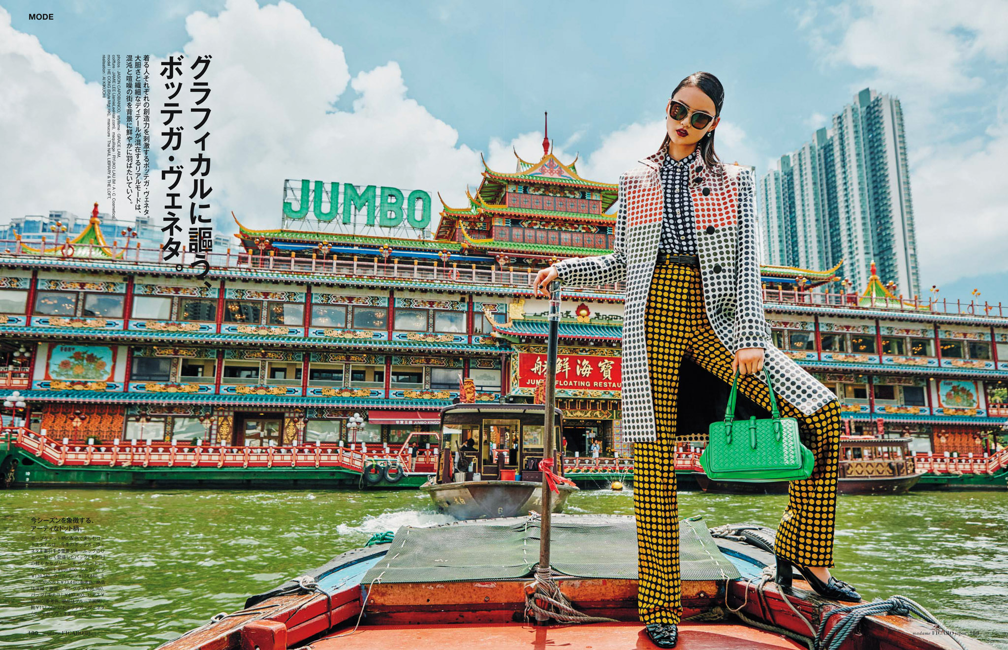 Woman poses in checkered coat and yellow pants in front of a large restaurant boat on the water against a city skyline backdrop.