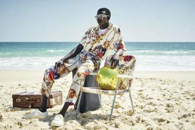 Man wearing patterned shirt and trousers sitting on folding chair on a sandy beach holding gold disco ball