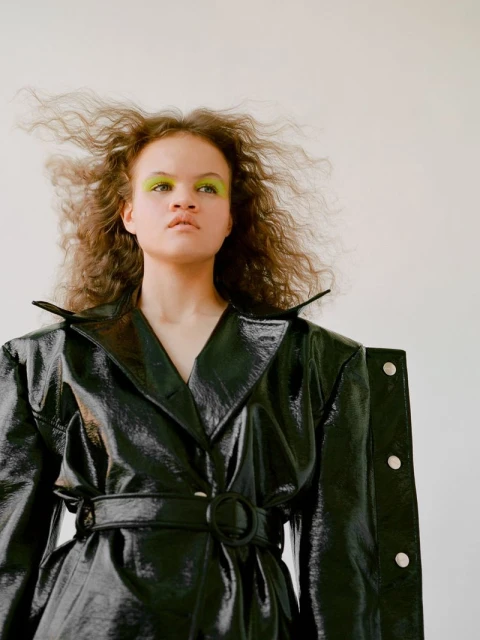 Woman in a glossy black trench coat with striking neon green makeup and wavy hair, posing indoors against a neutral backdrop.