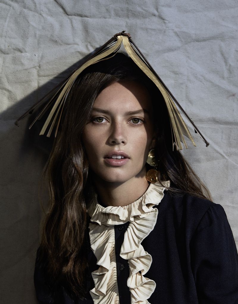 Brunette woman poses indoors with open book on head, wearing a ruffled-collar blouse and statement earrings against a textured backdrop.