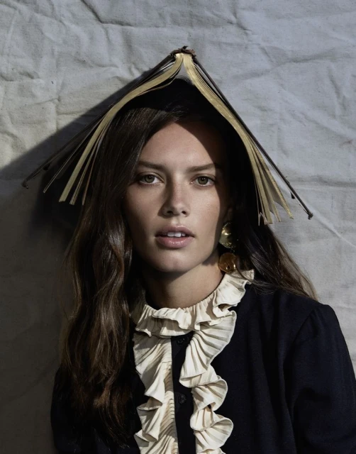 Brunette woman poses indoors with open book on head, wearing a ruffled-collar blouse and statement earrings against a textured backdrop.