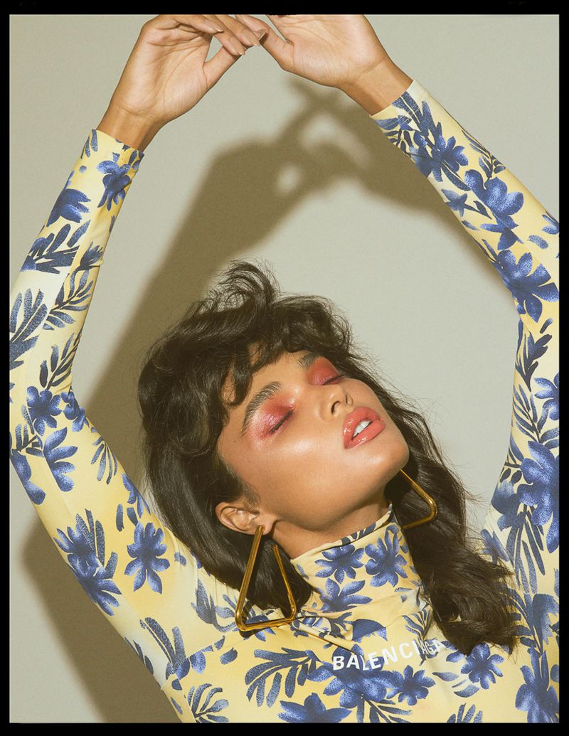Model with dark hair wearing a yellow and blue floral turtleneck with hands raised above head against a grey backdrop