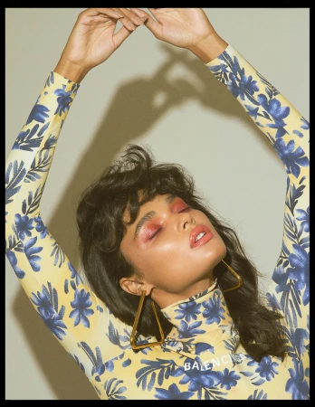 Model with dark hair wearing a yellow and blue floral turtleneck with hands raised above head against a grey backdrop