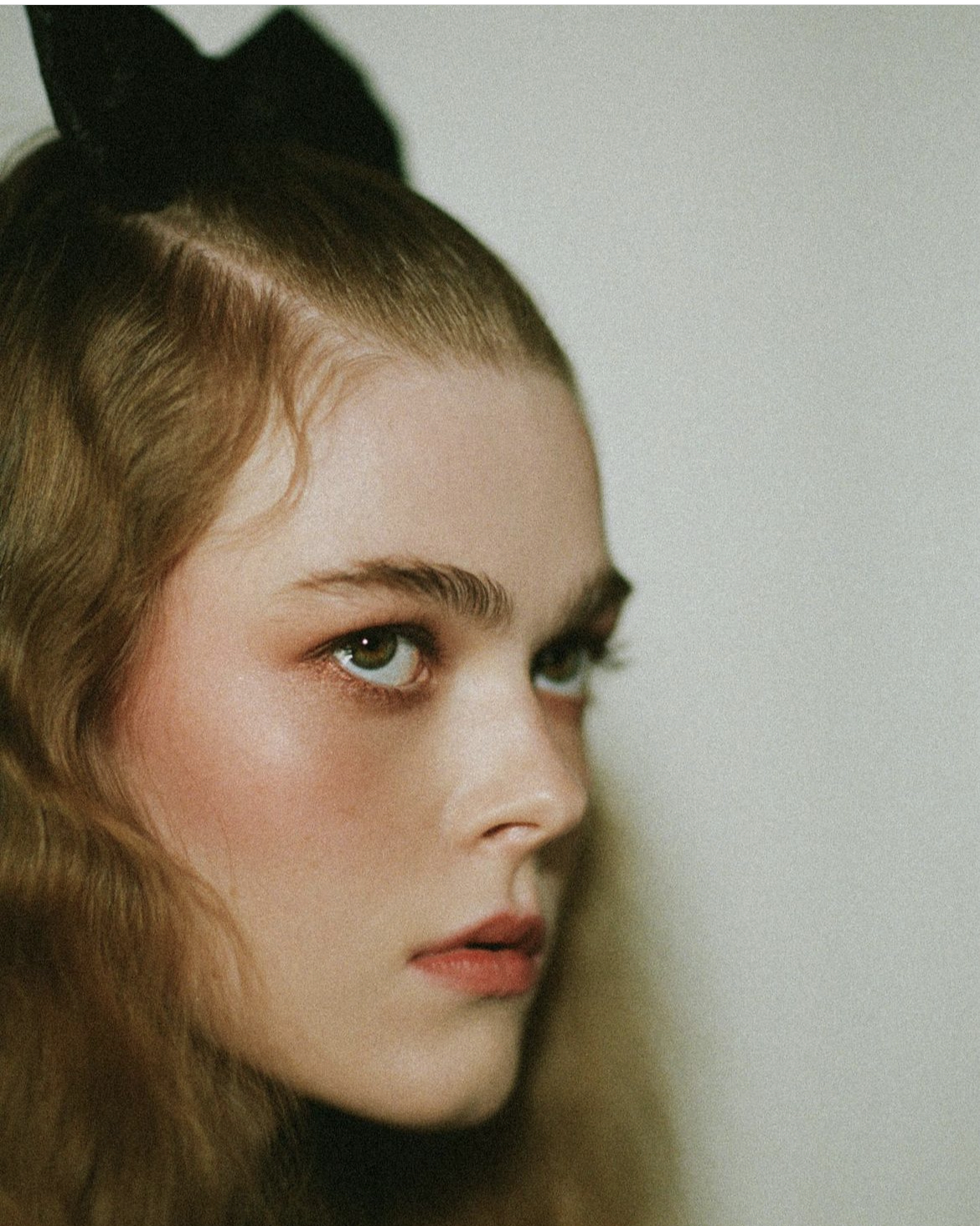Close-up portrait of a young adult woman with fair skin, soft makeup, and a black bow in her wavy hair, looking off to the side.