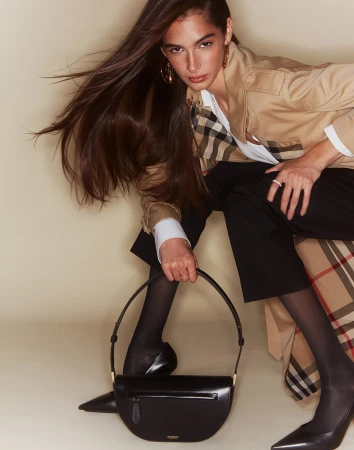 Model in camel trench coat holding a black handbag against a neutral studio backdrop with hair swept to the side.