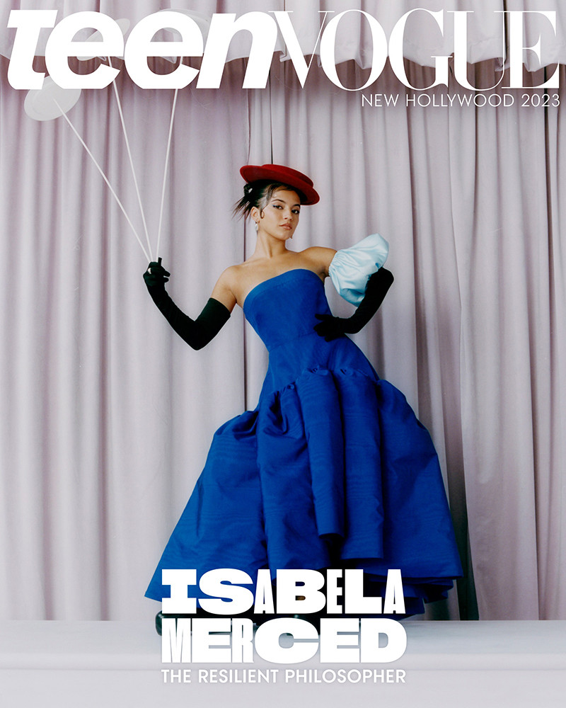 Isabela Merced on Teen Vogue cover, wearing a strapless blue dress with oversized puffed sleeves and a red hat, against a curtain backdrop.