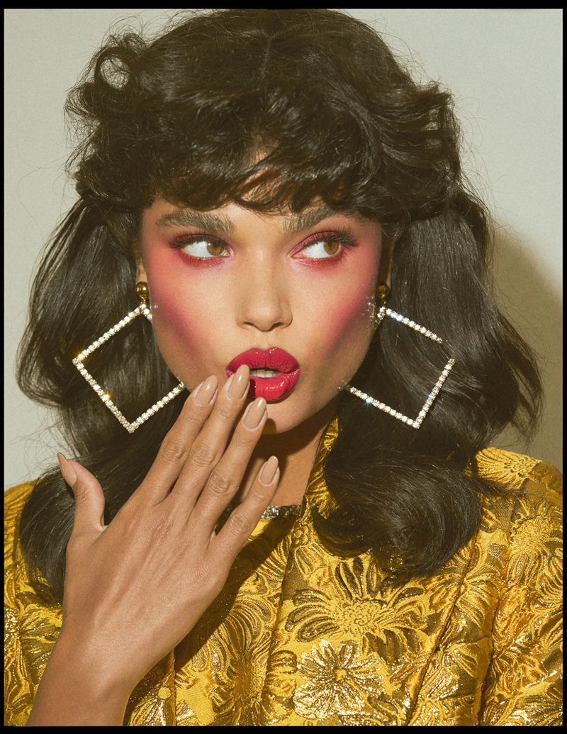 Portrait of a brunette woman with dramatic makeup and large, square, diamond earrings wearing a gold, floral brocade jacket.