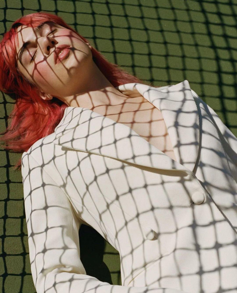 A person with red hair is lying down wearing a white blazer, with net shadow patterns cast across their face and clothing.