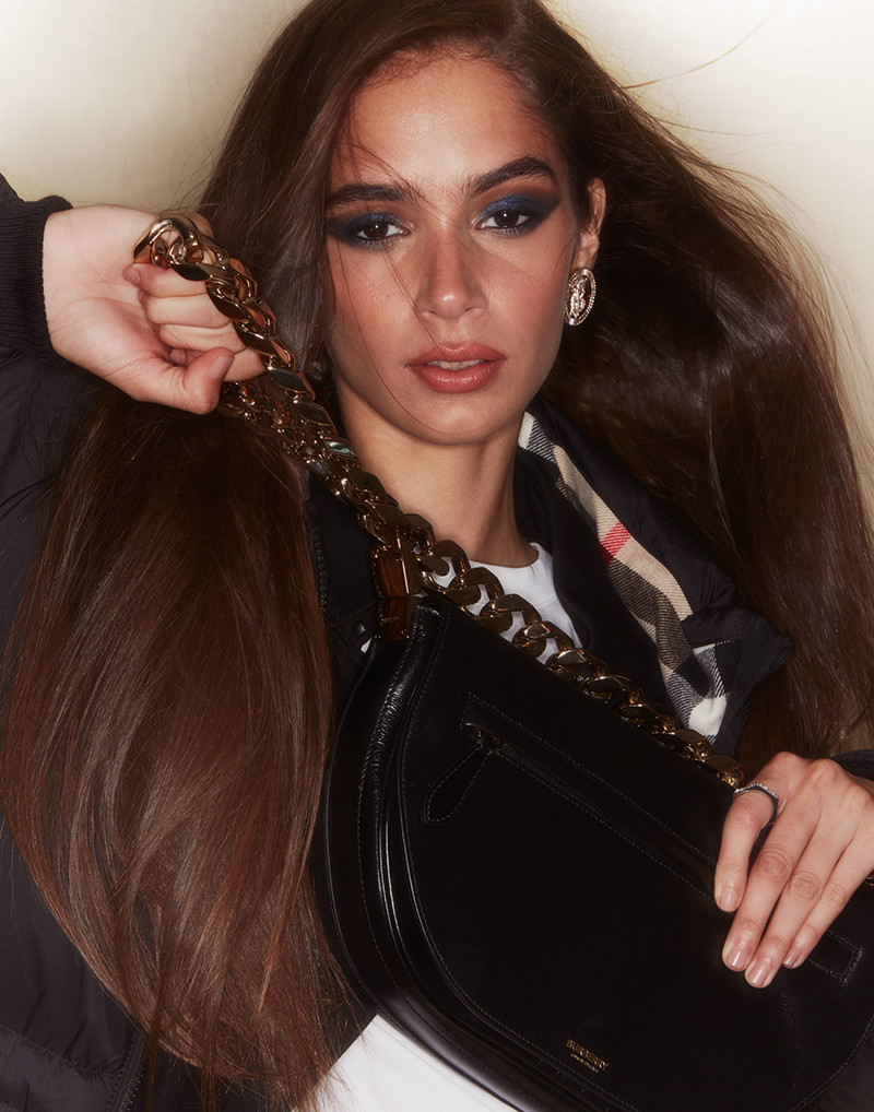 Close-up portrait of a long-haired brunette woman holding a black leather handbag with a chunky chain strap.