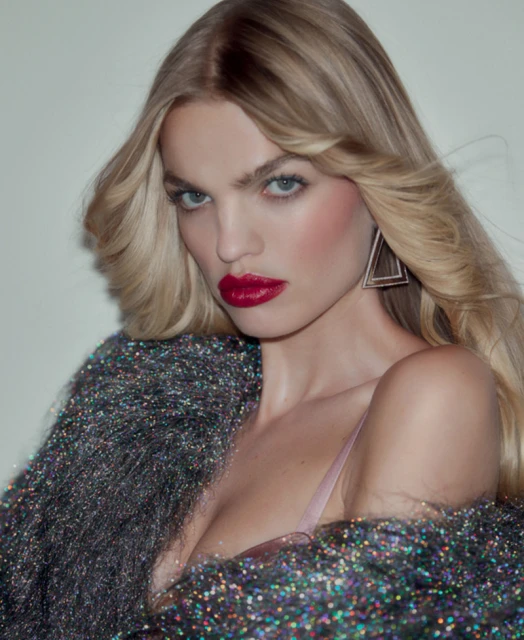 Blonde woman in shimmering jacket with red lipstick and triangle earrings posing in a close-up studio shot. Beauty and fashion.