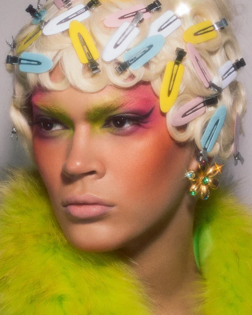 Close-up of a person with colorful makeup, blond curly hair, and hair clips, wearing a fluffy neon green jacket and sparkly earrings.