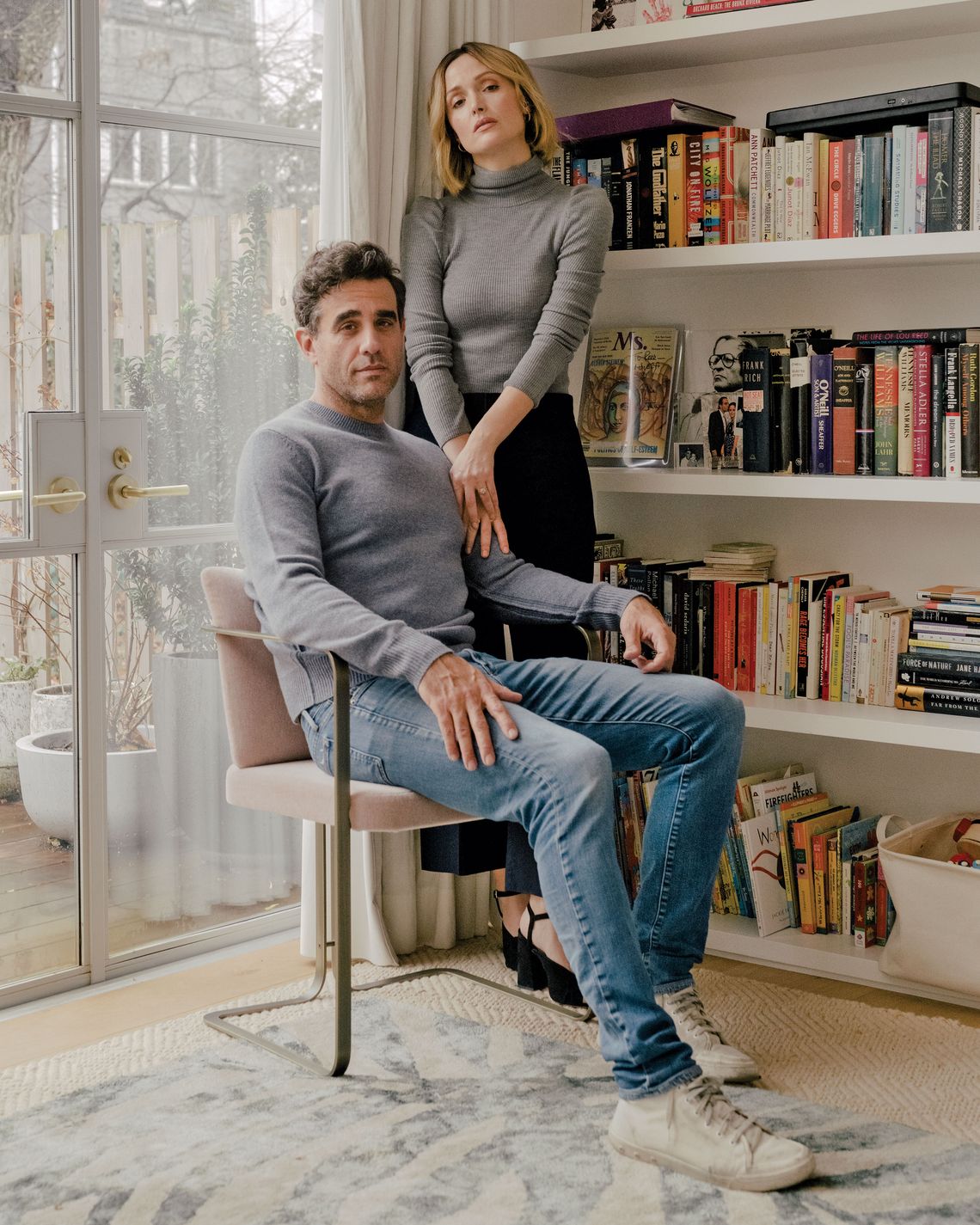 Indoor portrait of a Caucasian couple; a man sits in a chair, wearing jeans and a grey sweater; a woman stands next to him.